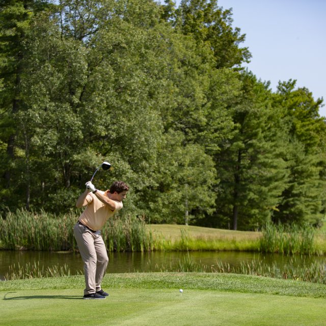 A golfer prepares to hit a ball over a stream, surrounded by green trees.