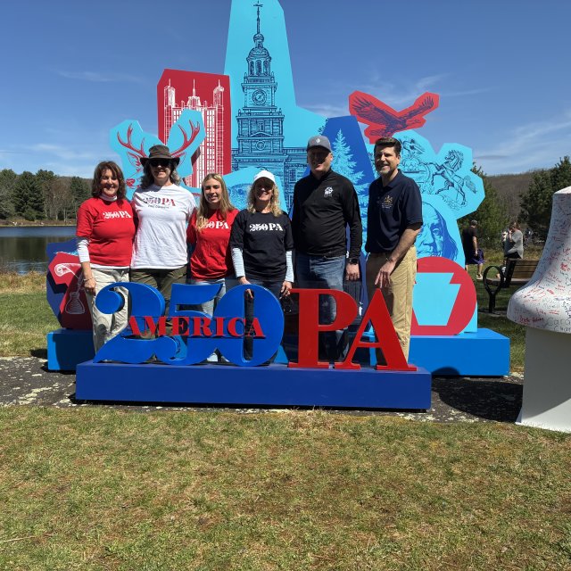 A group of people posing in front of an America 250PA exhibit.