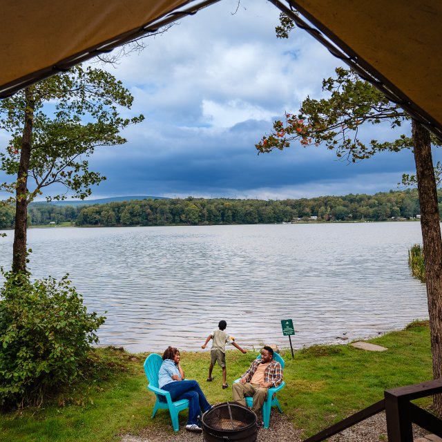 A couple talking around a firepit with their child skipping rocks in the lake behind them.