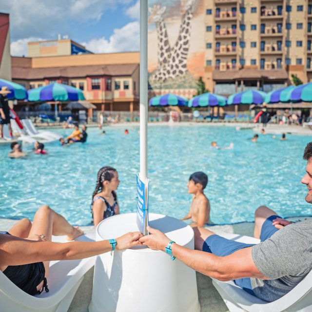 Mom and dad sit in beach chairs by a pool while their kids play in the water.