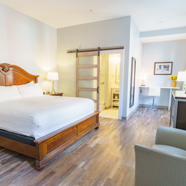 A large bed in a room with a chair, dresser, desk, TV and bathroom.