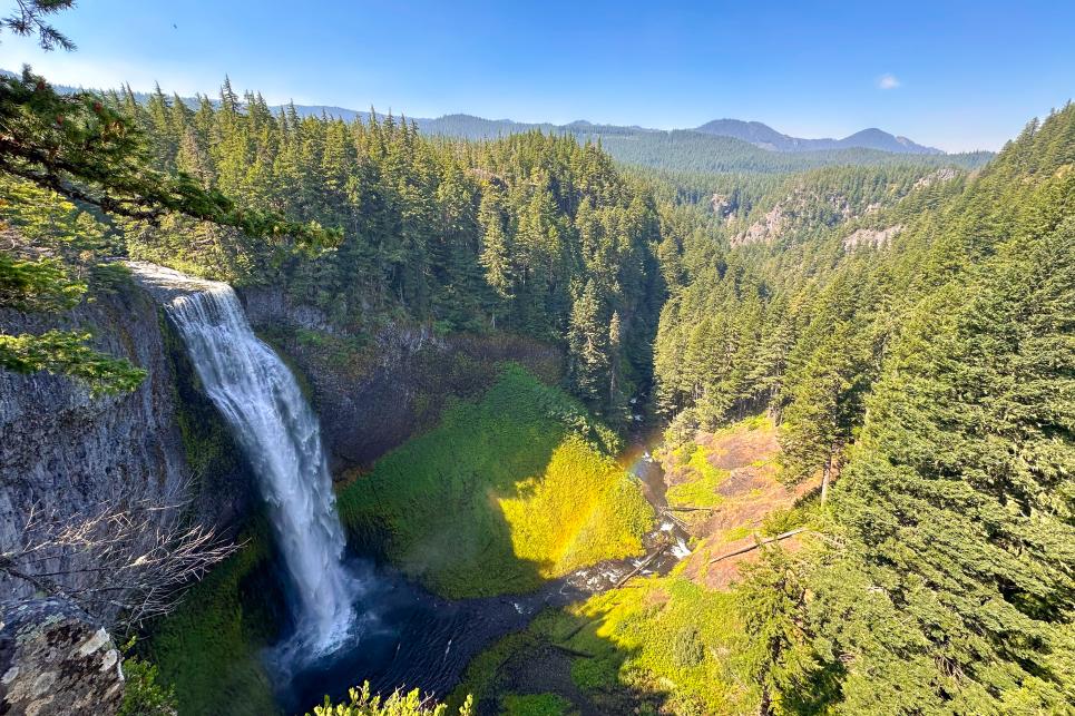 A view of a large waterfall in a vast forested canyon on a sunny day.