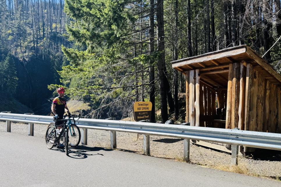 A cyclist stands by the guard rail on the Aufderheide with the wood structure at the foot of the trailhead to the hot springs.