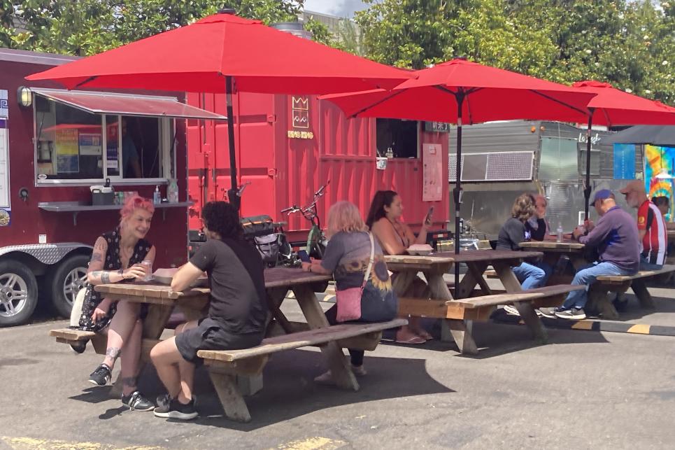 Diners sit under red umbrellas at picnic tables placed in the parking lot with food trucks.