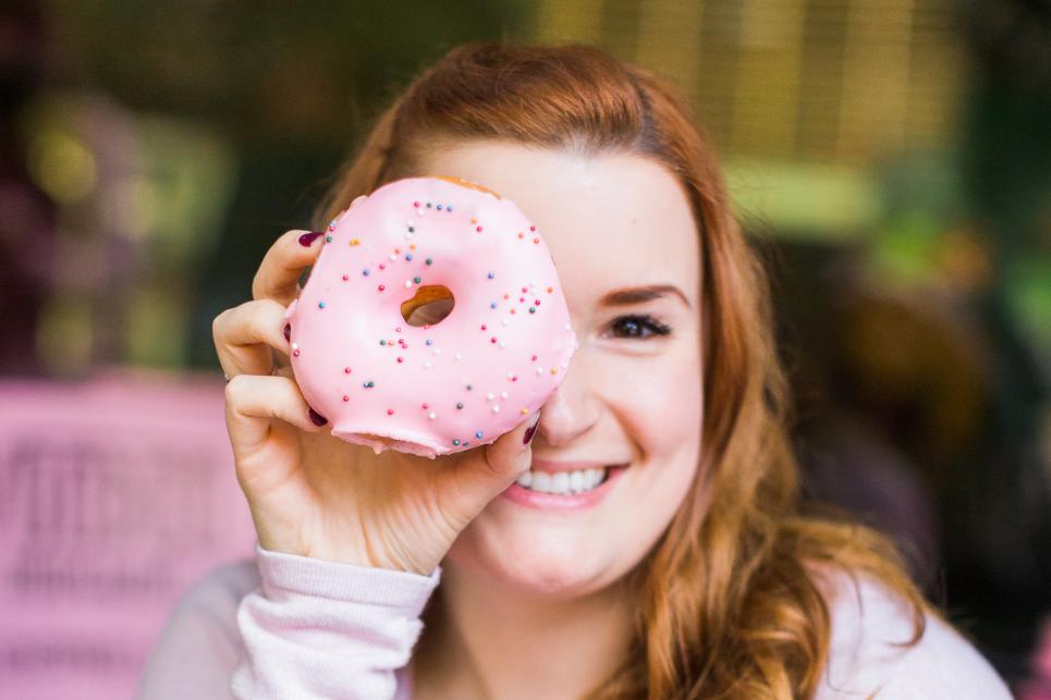 A person holds a pink glazed doughnut up looking through the whole with one eye.