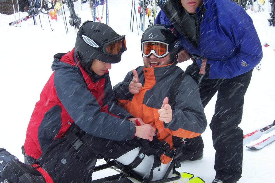 Two skiers help a skier strap onto an adaptive skiing board. They are on the snow and snow is falling.