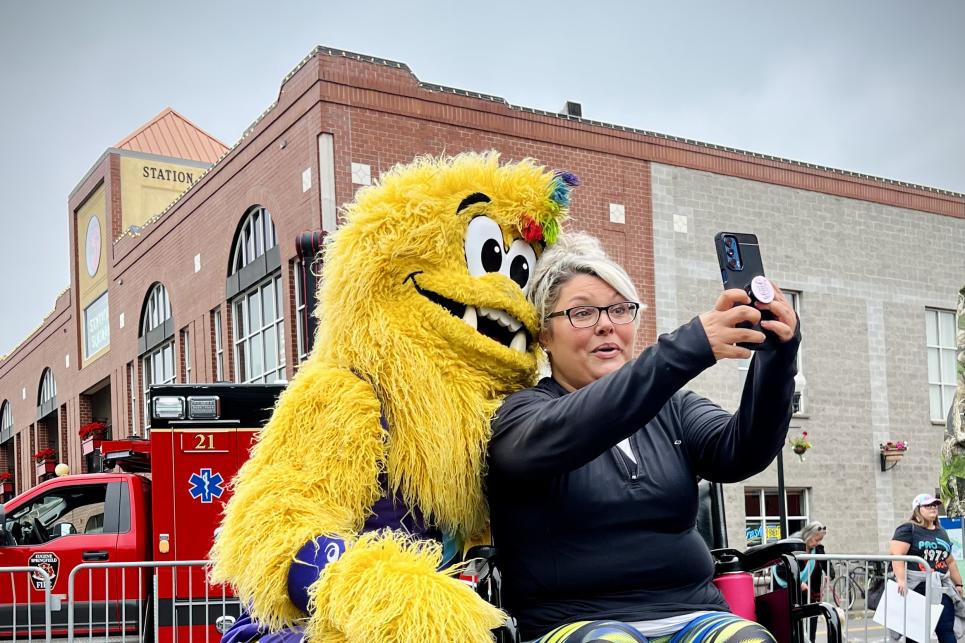 A woman with a wheelchair in running gear takes a selfie with the World Athletics Championships' Legend — a yellow Big Foot mascot wearing running shoes.