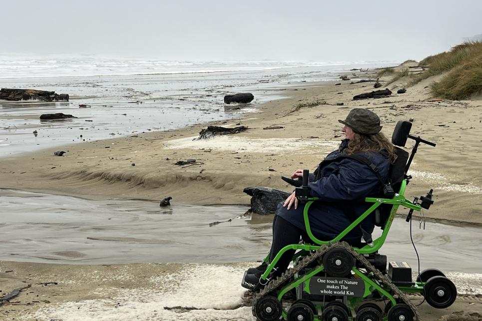 A person sits in a green track chair on the sandy beach next to a small stream.