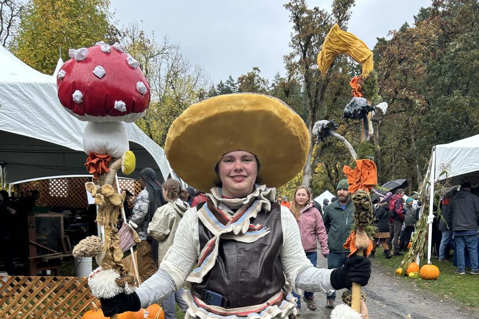 A person stands dressed as a mushroom holding two decorated staffs with mushrooms on them.