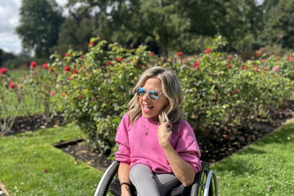 Kristin in a pink shirt sits in her wheelchair by the roses.