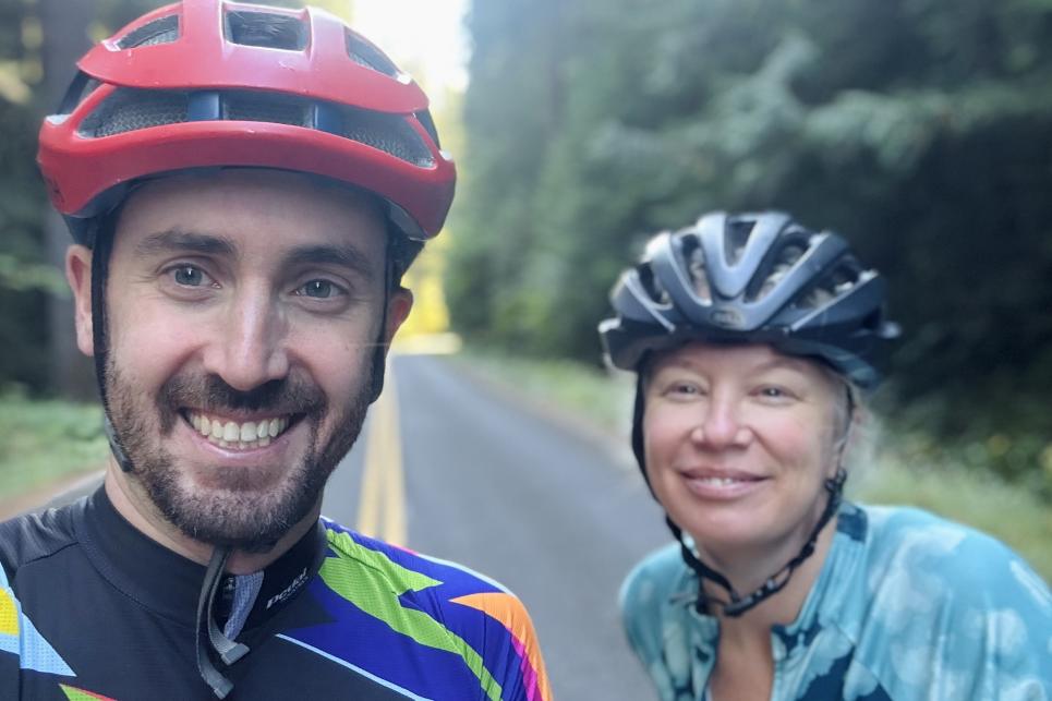 Two people in bicycle helmets pose on the roadside.