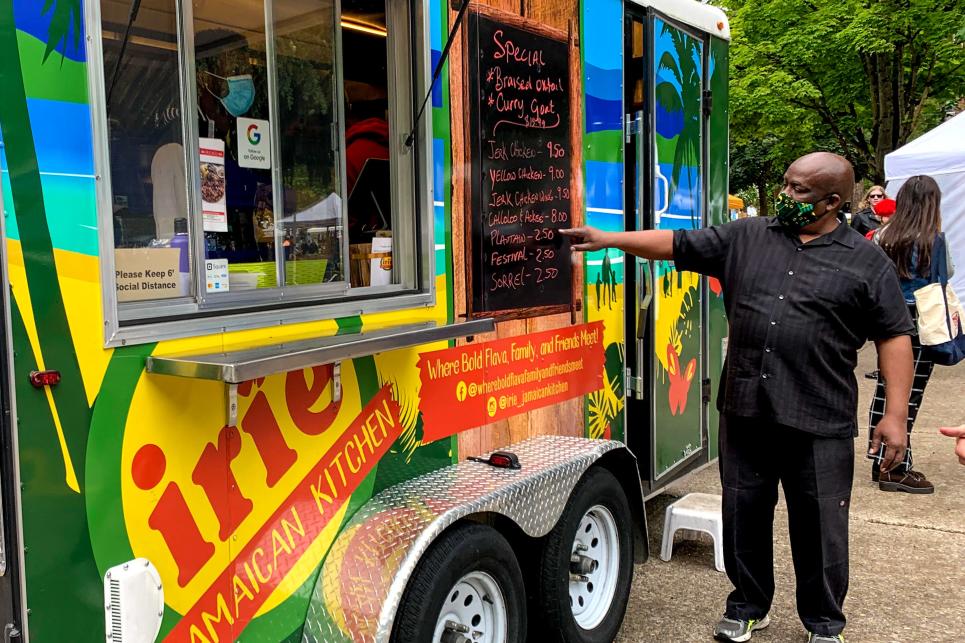 a man stands in front of a food truck pointing at the menu which features Jamaican dishes.
