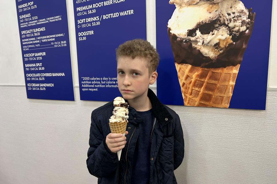 A young kid holds a waffle cone with three scoops of vanilla and chocolate ribboned ice cream stacked high.