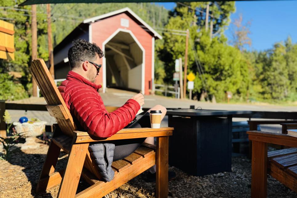 A person in a red jacket sits in a wood chair with coffee. The red Office Bridge is in the background.