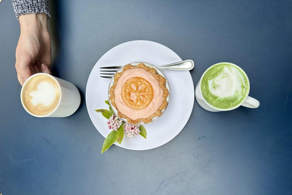 A latte, a decadent mini pie and a matcha latte line up in a row on a blue table. A hand reaches in for the first drink.