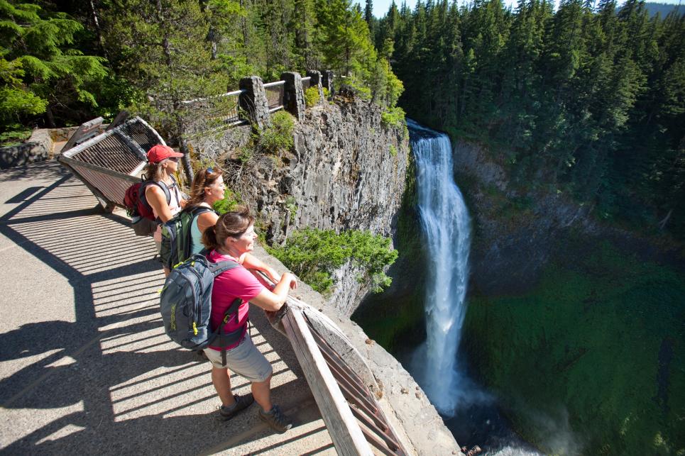 Three women with backpacks and warm weather clothing lean against the railing overlooking Salt Creek Falls.
