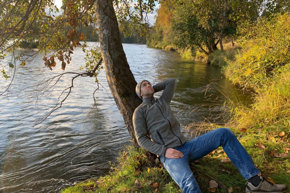 Man sits leaning against a tree beside the Willamette River with fall foliage surrounding him.