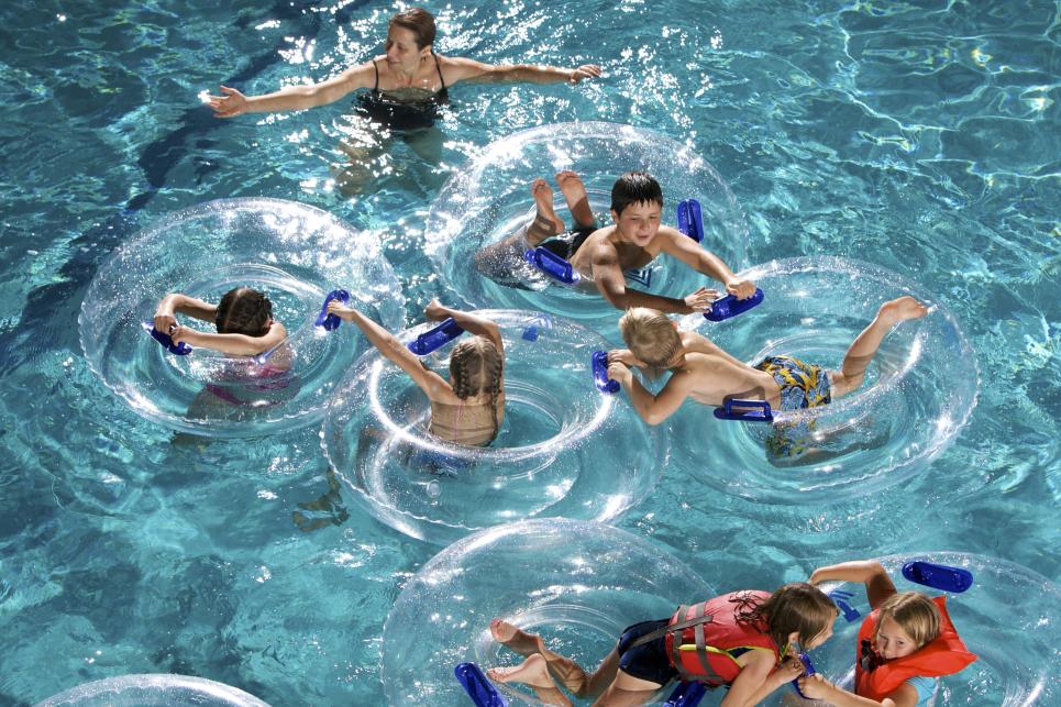 Children in clear plastic inner tubes form a circle in the pool. A woman and other children in life jackets play in the water nearby.