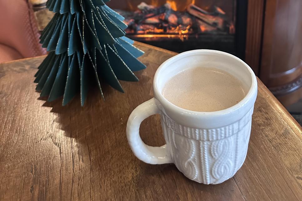 A white sweater patterned coffee cup with a warm beverage sitting on a table next to a gas fireplace.