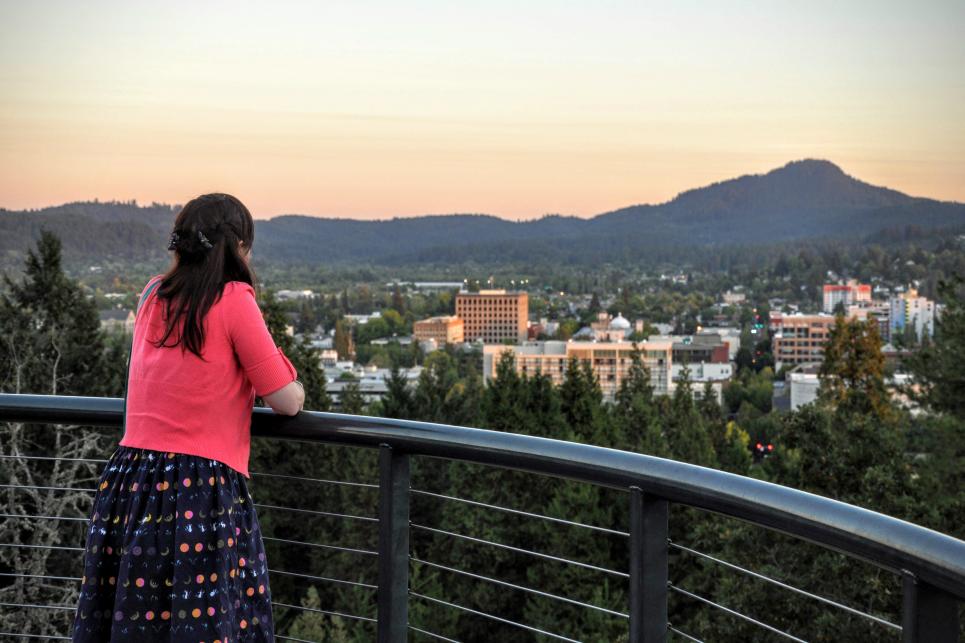 A woman stands at railing overlooking the city of Eugene with Spencer Butte in the background.