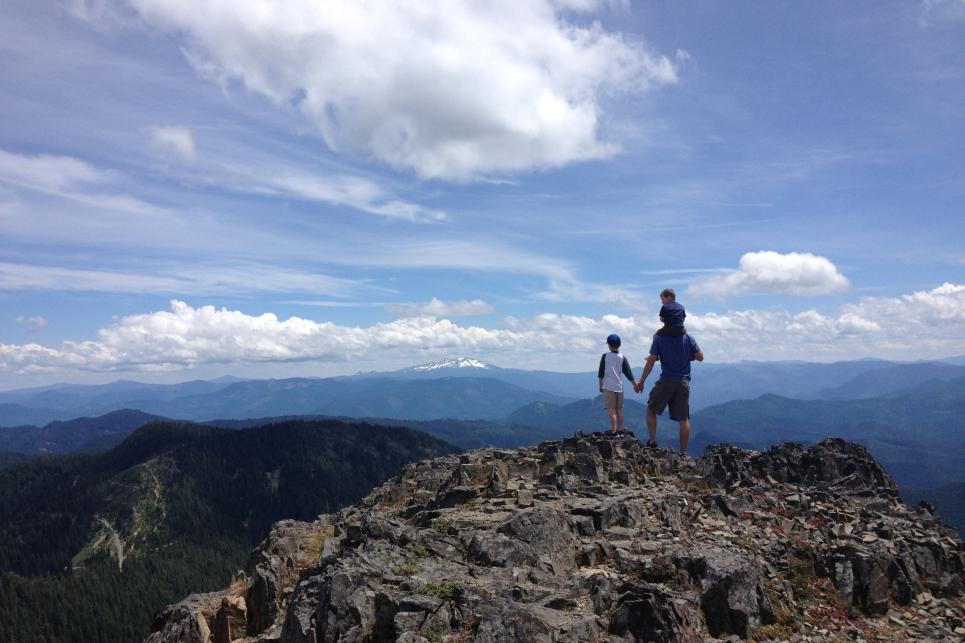 A family holding hands, standing at the top of a rocky mountain overlooking distant hills below.