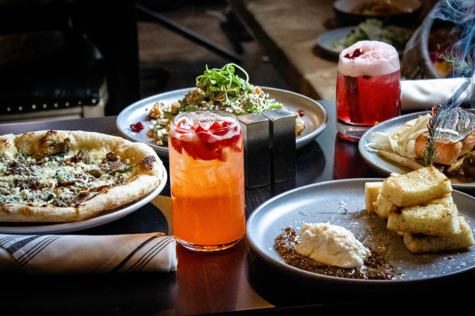 An array of food and drink on a table top that includes pizza and some colorful cocktails.
