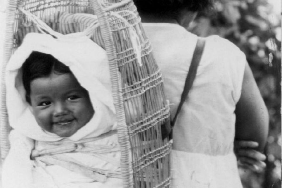 An Indigenous Siletz woman carries a smiling baby on her back in a woven cradleboard. Photo is black and white.