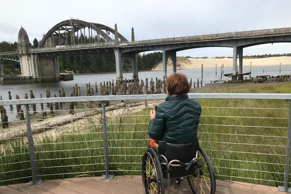 A person in a dark puffy coat with a wheelchair is on the flat boarded viewing point looking out toward the historic Siuslaw River Bridge. The viewing platform has a thin rail and wire fence allowing optimal viewing with the top rail below the person's sightline. Green rushes and grass fill in the space between the deck and riverside. Sand dunes are in the background.