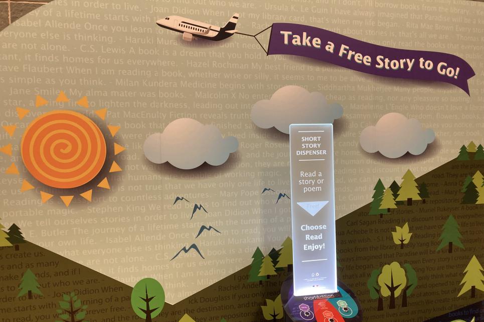 A colorful backdrop with forest, sun, clouds and plane with a banner that reads "Take a Free Story to Go!" banner behind a kiosk.