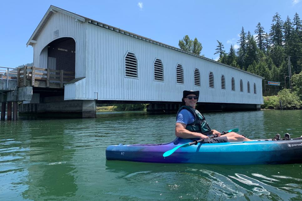 Man in life jacket in blue kayak pauses on the water in front of the white covered bridge.