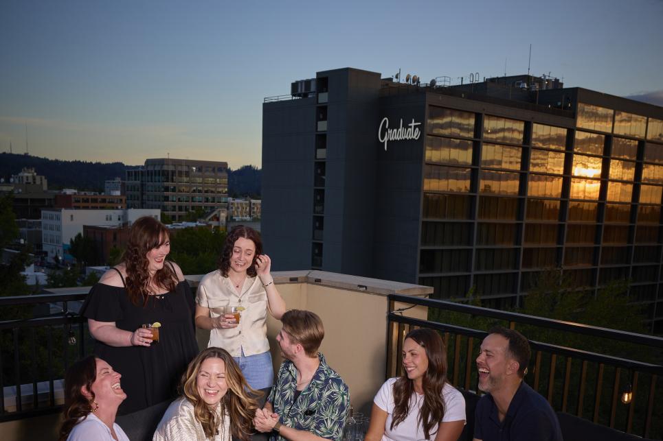 A group gathers around the fire on the rooftop deck with the sun sinking in the background.
