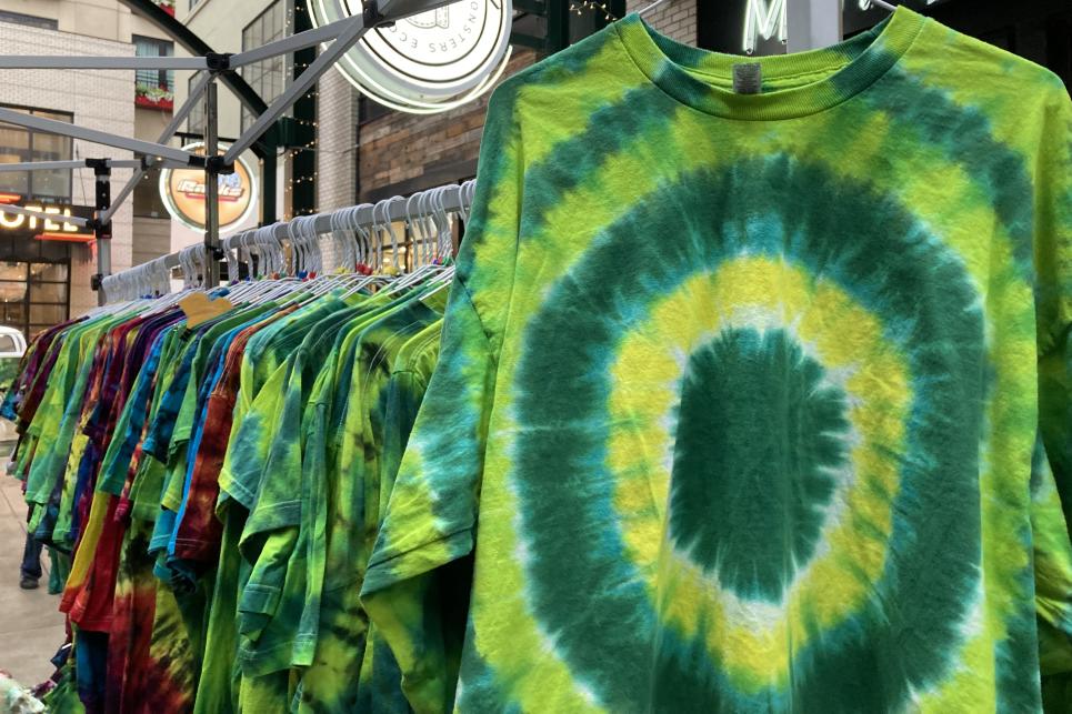 A yellow and green tie-dye shirt with a circle pattern hangs in front of a rack of more t-shirts.