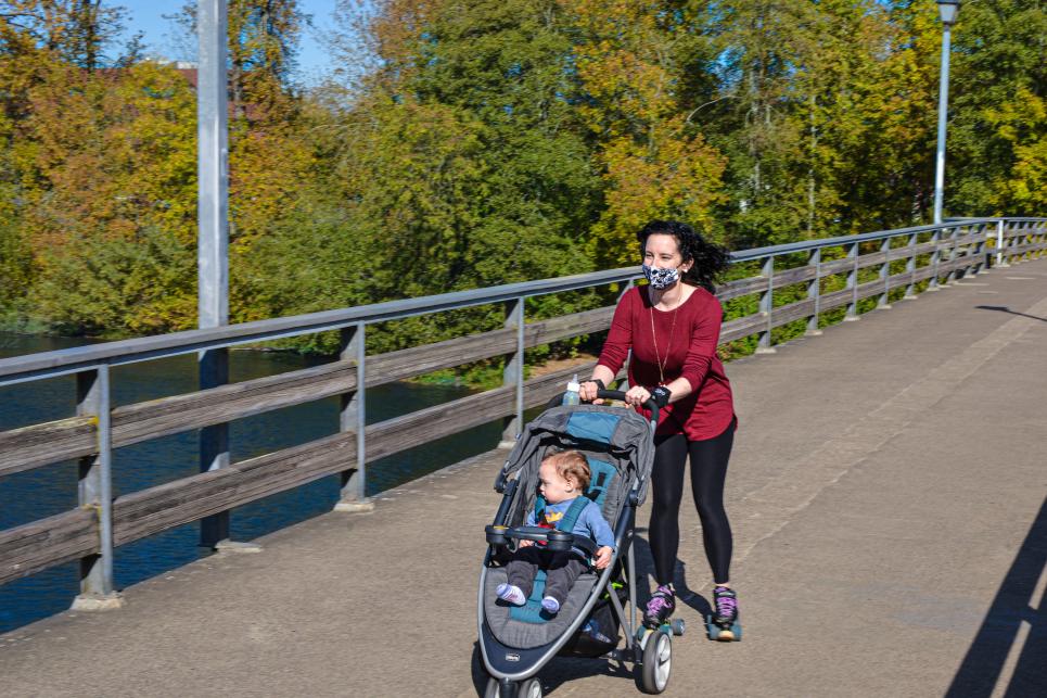 A woman in a red top and face mask roller-skates pushing a jogging stroller with a child across a pedestrian bridge. The bridge is paved and smooth with rails. Fall colored trees and river are in the background.