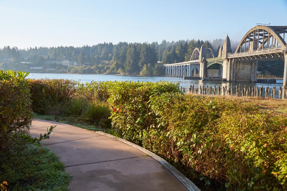 A walking path winds toward the Siuslaw River with the Highway 101 bridge in the background.
