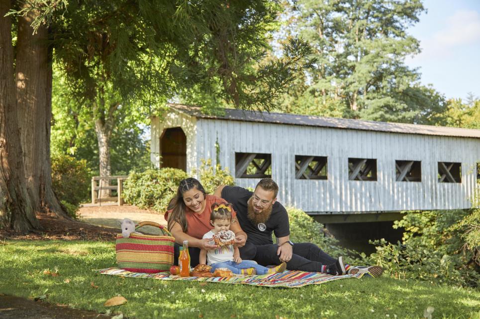 A family of three enjoys a picnic of pastries at a park with a white covered bridge in the background.