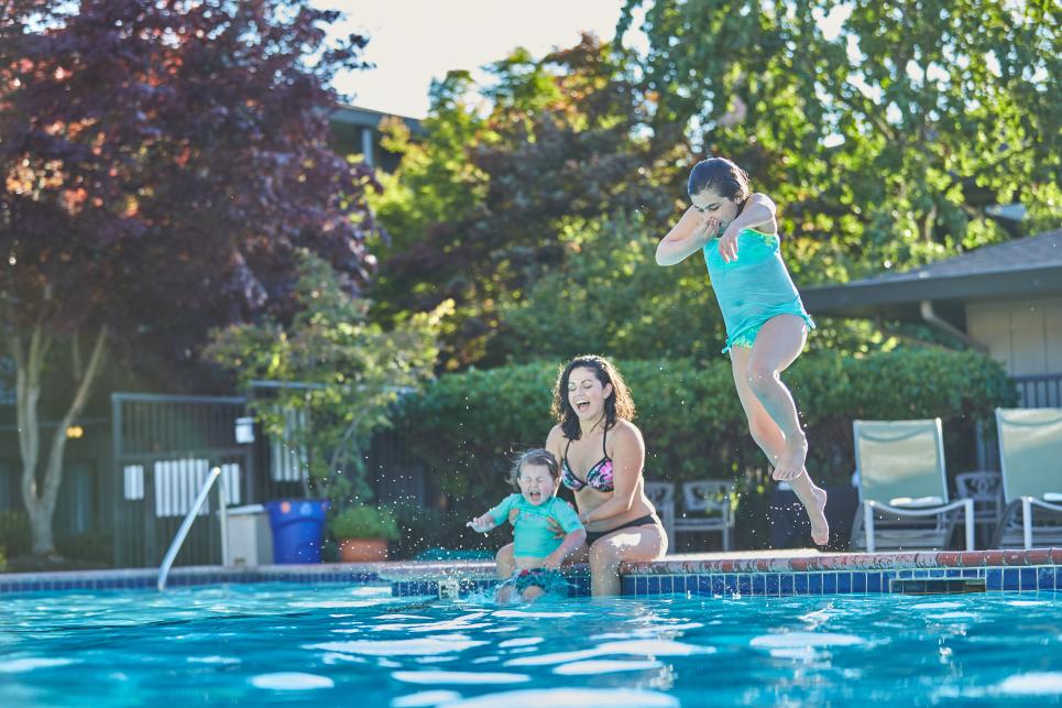 A mom with a baby sits on the edge of the pool while a child jumps in.