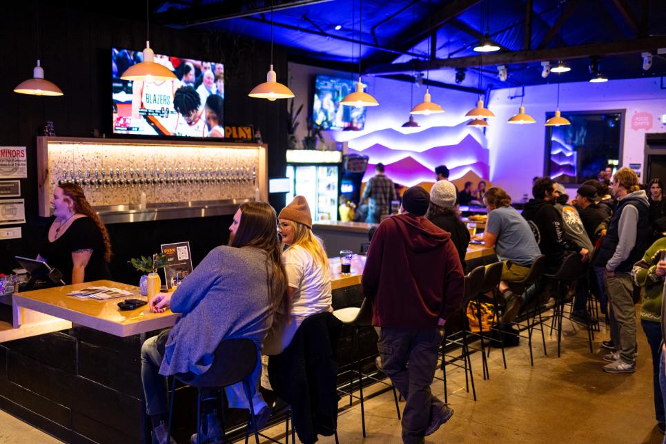 Young people sit and stand around the bar with dim lighting and a large screen TV.