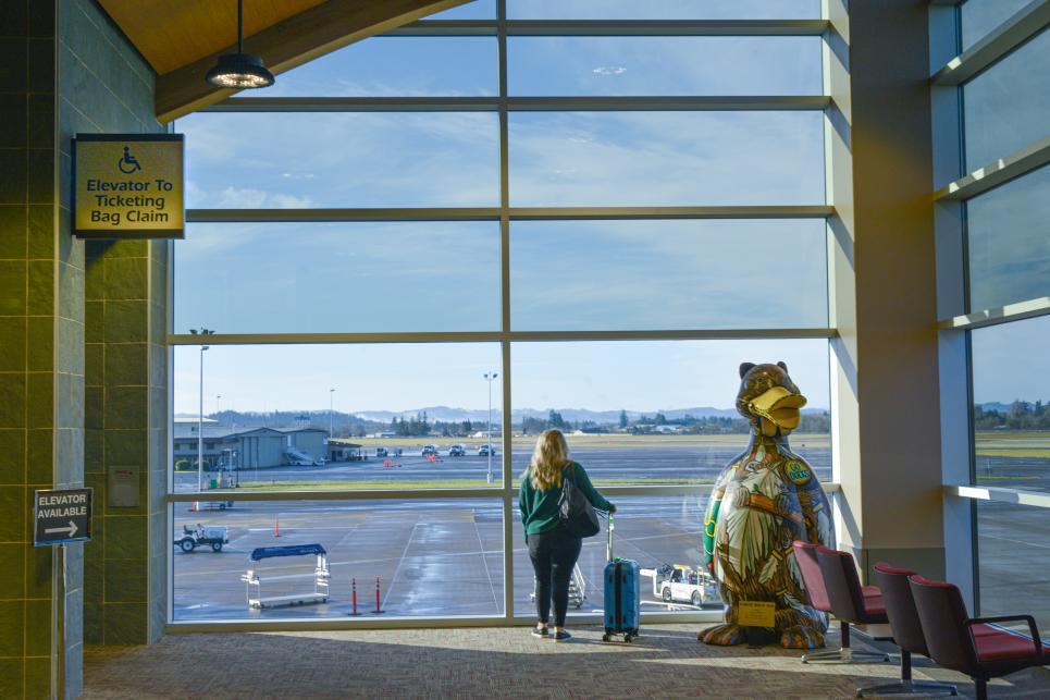 A woman with a rolling carry on suitcase stands in front of large windows at the airport looking out over the tarmac.