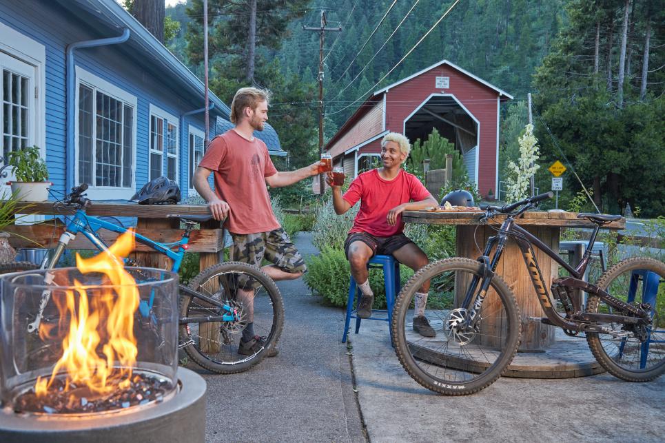Two mountain bikers with their bikes nearby sit and cheers with their beers on a patio at Westfir Lodge in the evening. There is a fire in a firepit in the foreground and the red Office Covered Bridge in the background