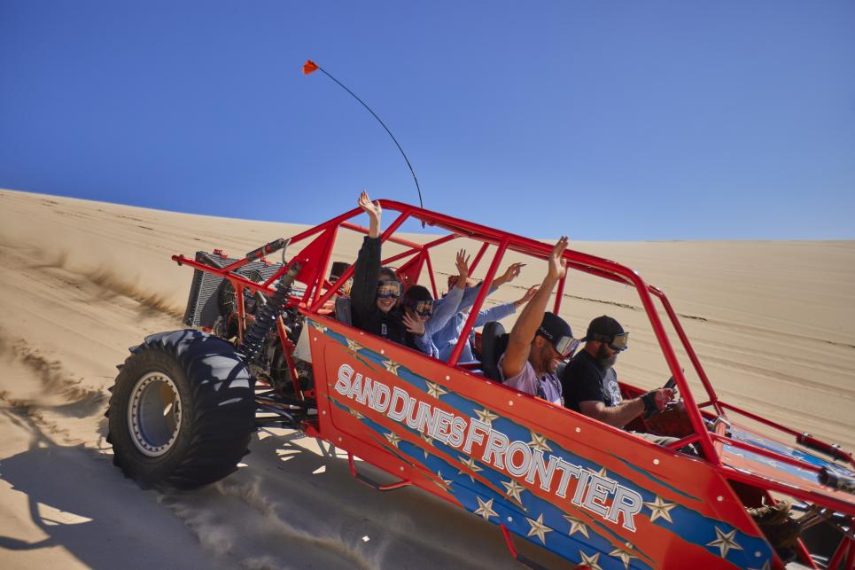 Group of friends raise their hands in a dune buggy that's driving down the dunes at a high speed.