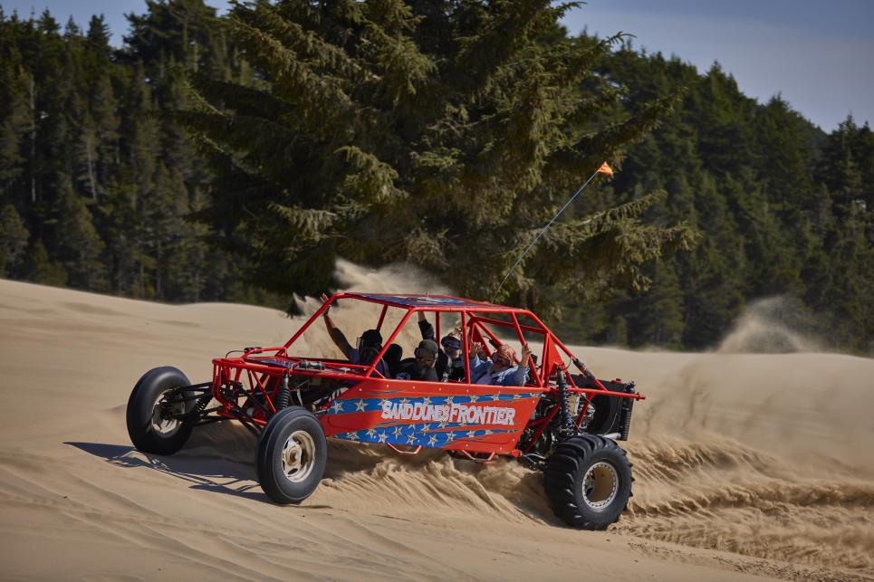 A Dune buggy full of people with their arms in the air and smiles on their faces turns in the sand causing sand to fly in the air.