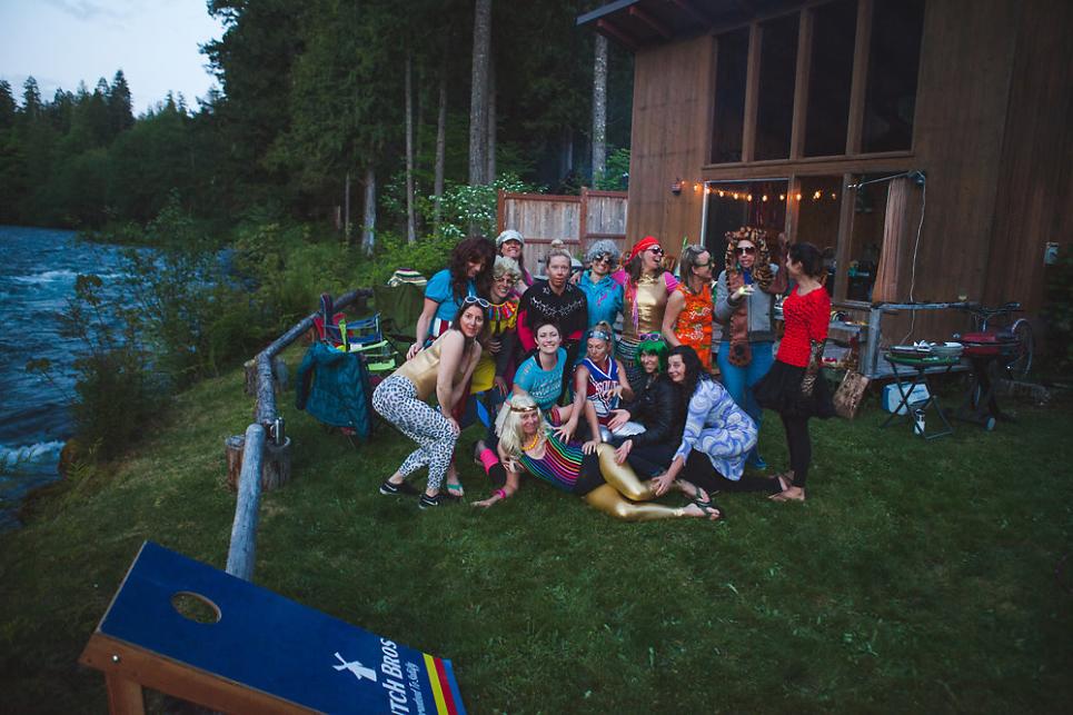 A group of friends in silly costumes pose for a photo between a cabin and the river
