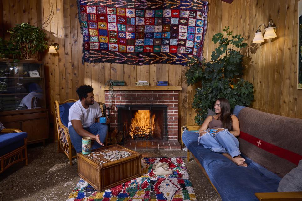 People sit in the wood paneled living area by a roaring fireplace with a puzzle and a warm beverage.