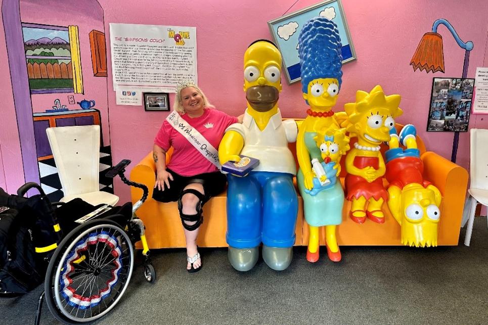 Melinda in her Ms. Wheelchair sash sits next to Homer on The Simpsons' couch.