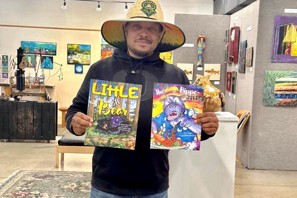 Andre Royal holds up two children's books. The gallery's art work is behind him.