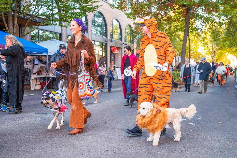 Two costumed people walk their costumed dogs in a pet parade downtown.
