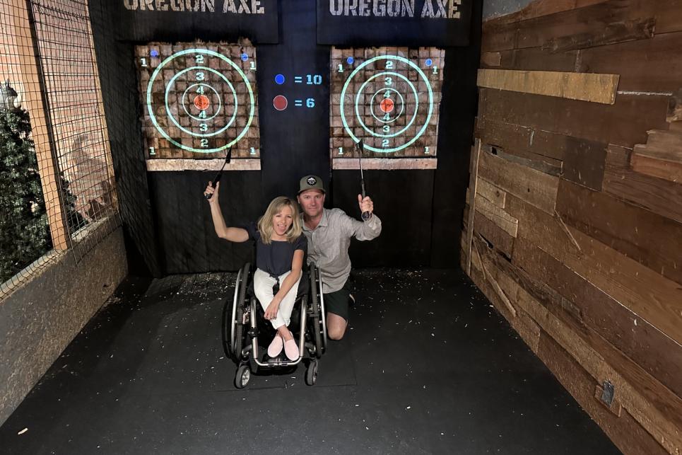 Kristin, a petite blond woman using an wheelchair, with her husband, each holding up an axe, pose in front of the targets at Oregon Axe.