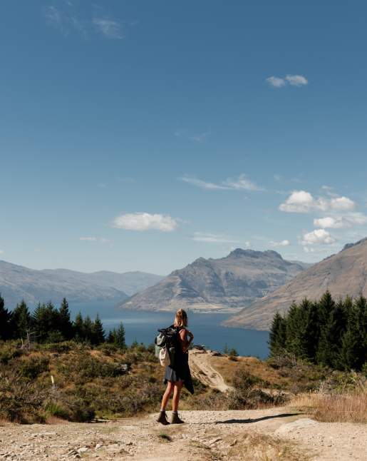 A girl standing at the top of Queenstown Hill after hiking up. She is looking out to the view of the town, Lake Whakatipu and mountain ranges.