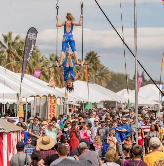 Large crowd outdoors between white tent booths. Two acrobats on a bar hang over the crowd