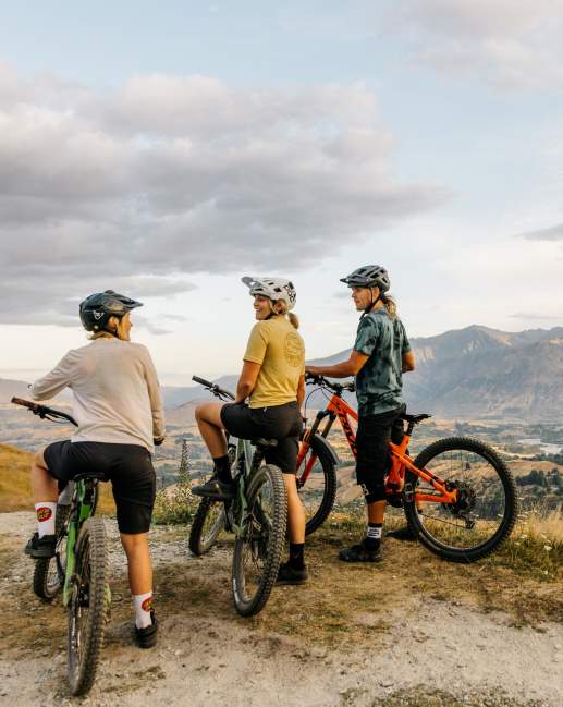 Three mountain bikers at the top of a mountain overlooking a valley with the Remarkables mountains in the background